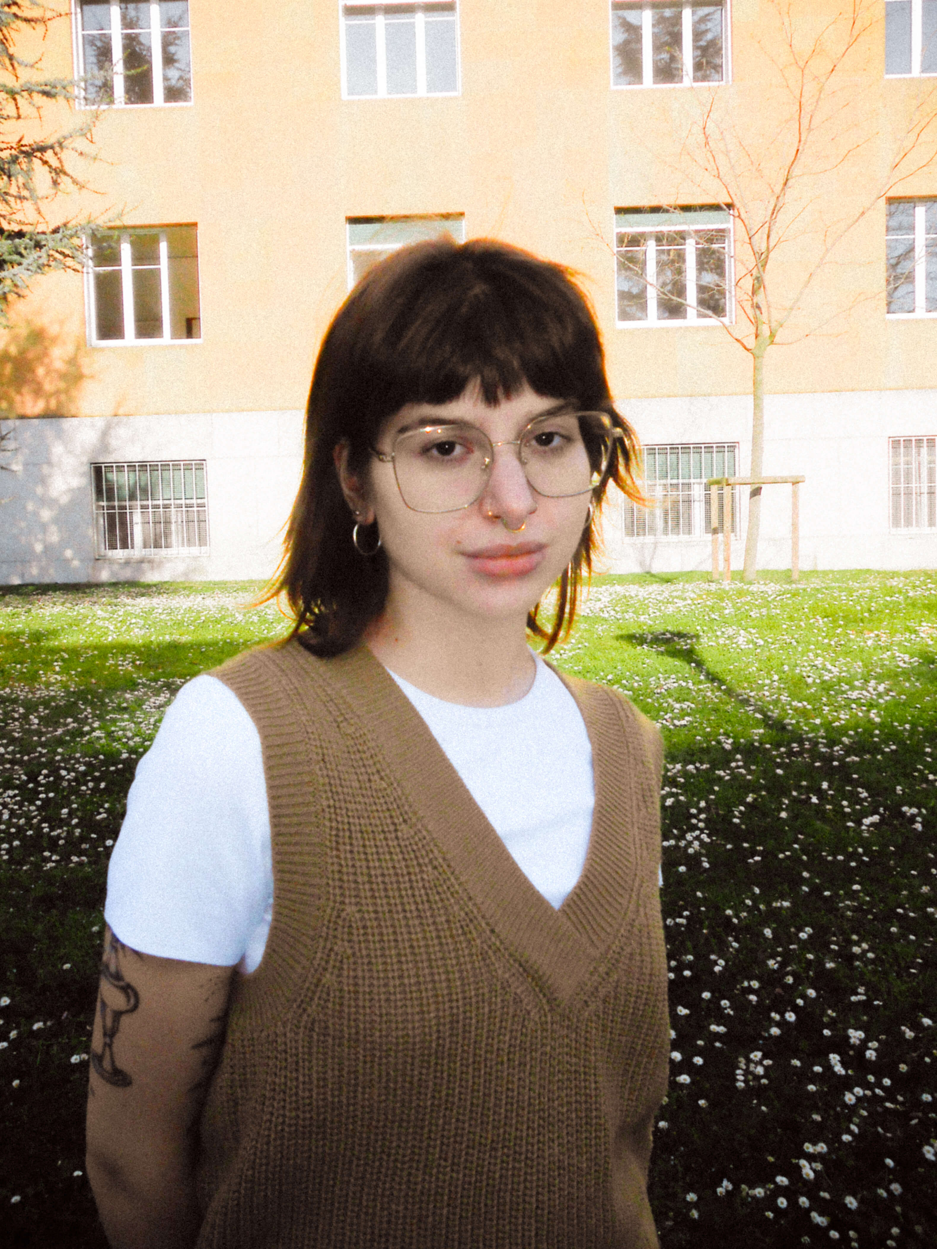 Portrait d’une jeune femme au style affirmé, debout en extérieur devant un bâtiment moderne. Elle porte un t-shirt blanc sous un gilet en maille marron, des lunettes rondes, des anneaux aux oreilles et un piercing au nez. Un tatouage est visible sur son bras gauche. L’arrière-plan montre une pelouse fleurie et un arbre sans feuilles, suggérant une saison intermédiaire.