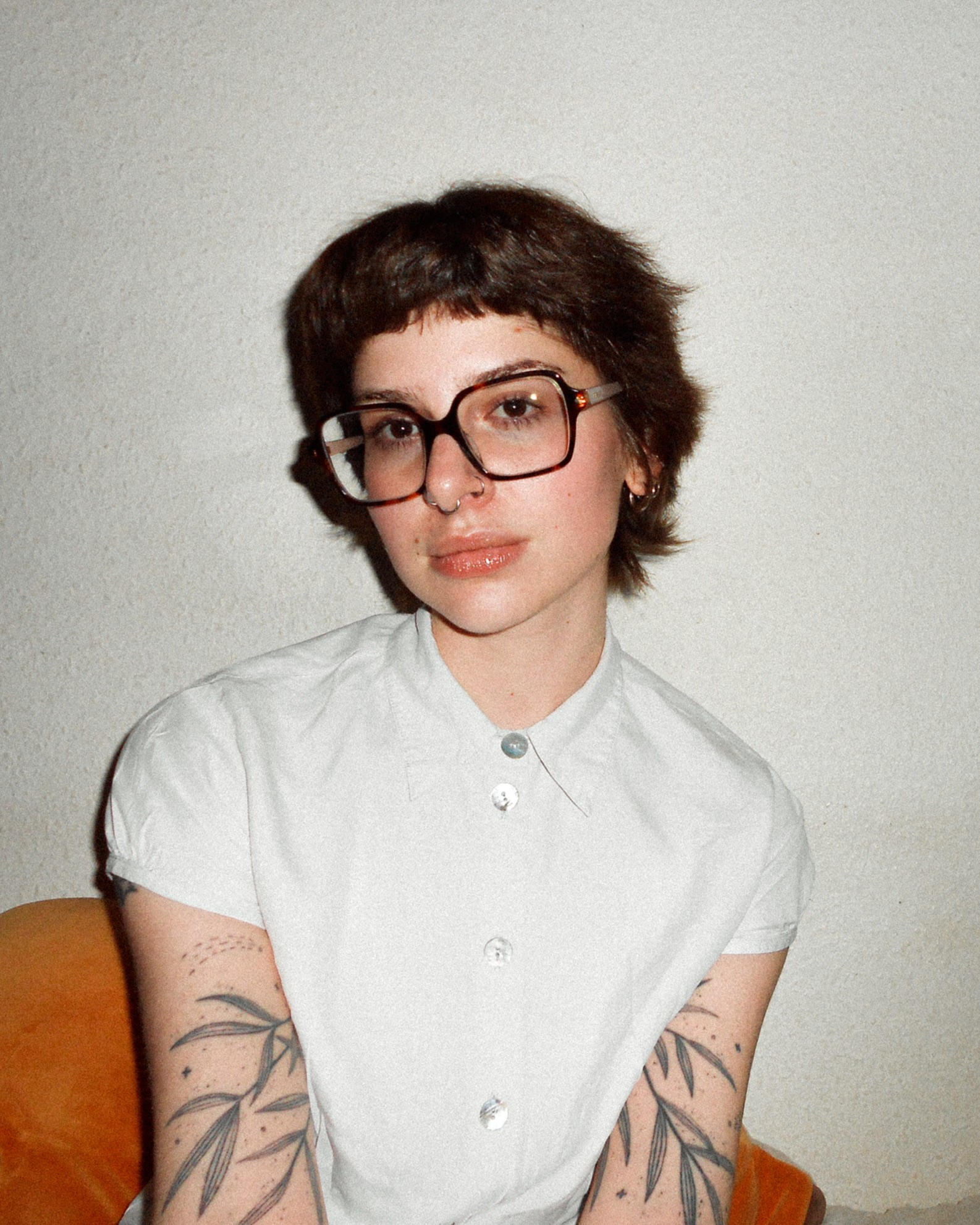 Portrait d’une jeune femme au style affirmé, debout en extérieur devant un bâtiment moderne. Elle porte un t-shirt blanc sous un gilet en maille marron, des lunettes rondes, des anneaux aux oreilles et un piercing au nez. Un tatouage est visible sur son bras gauche. L’arrière-plan montre une pelouse fleurie et un arbre sans feuilles, suggérant une saison intermédiaire.
